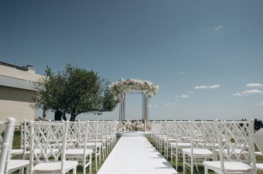 Lüks bir düğün sahnesi, beyaz bir patika boyunca dizilmiş beyaz dantel sandalyeli yüksek çiçekli bir kemer.