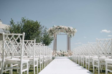 Lüks bir düğün sahnesi, beyaz bir patika boyunca dizilmiş beyaz dantel sandalyeli yüksek çiçekli bir kemer.