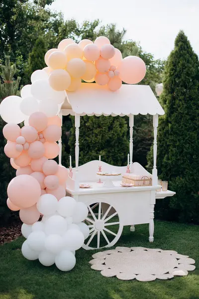 Tekerlekleri olan beyaz ahşap bir araba ve yemyeşil bir bahçede balonlarla süslenmiş şeker kalıbı görevi gören bir kubbe. Düğünler ve doğum günleri için mükemmel.