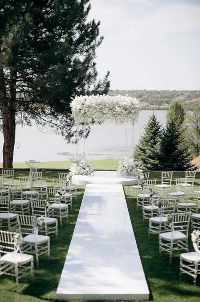 Göl kenarında beyazlarla süslenmiş lüks bir açık hava düğün töreni. Zarif bir çiçek kemeri, şeffaf sandalyeler, yeşil bir çimen ve fevkalade bir arka plan.