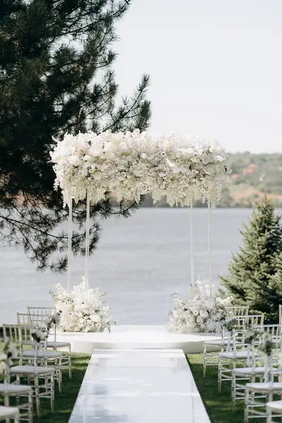 Göl kenarında beyazlarla süslenmiş lüks bir açık hava düğün töreni. Zarif bir çiçek kemeri, şeffaf sandalyeler, yeşil bir çimen ve fevkalade bir arka plan.