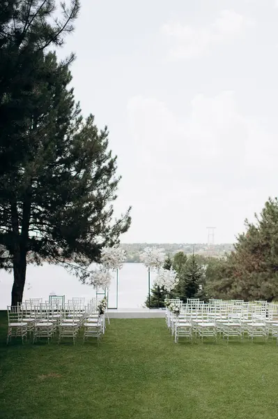 Göl kenarında beyazlarla süslenmiş lüks bir açık hava düğün töreni. Zarif bir çiçek kemeri, şeffaf sandalyeler, yeşil bir çimen ve fevkalade bir arka plan.