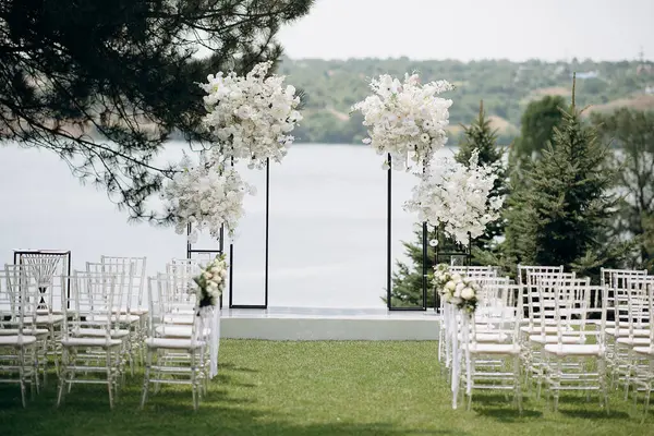 Göl kenarında beyazlarla süslenmiş lüks bir açık hava düğün töreni. Zarif bir çiçek kemeri, şeffaf sandalyeler, yeşil bir çimen ve fevkalade bir arka plan.