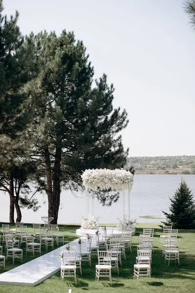 Göl kenarında beyazlarla süslenmiş lüks bir açık hava düğün töreni. Zarif bir çiçek kemeri, şeffaf sandalyeler, yeşil bir çimen ve fevkalade bir arka plan.