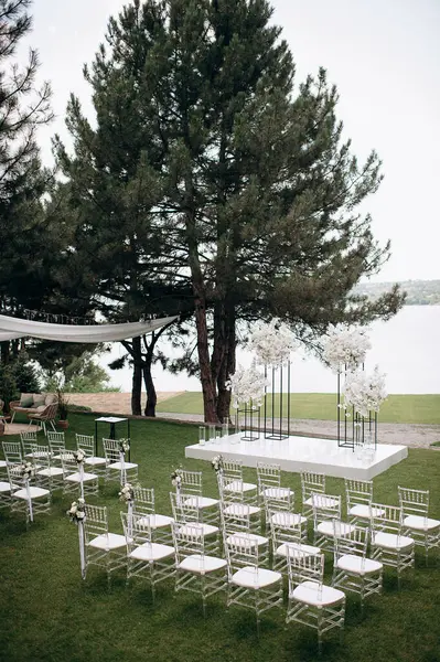 Göl kenarında beyazlarla süslenmiş lüks bir açık hava düğün töreni. Zarif bir çiçek kemeri, şeffaf sandalyeler, yeşil bir çimen ve fevkalade bir arka plan.
