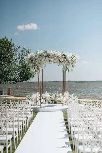 Göl kenarında beyazlarla süslenmiş lüks bir açık hava düğün töreni. Zarif bir çiçek kemeri, şeffaf sandalyeler, yeşil bir çimen ve fevkalade bir arka plan.