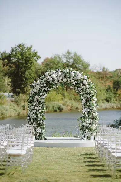Nehir kenarında çiçek kemeri ve şeffaf sandalyeleri olan güzel bir açık hava düğün töreni. Zarif beyaz ve yeşil dekor doğal ile harmanlanır.