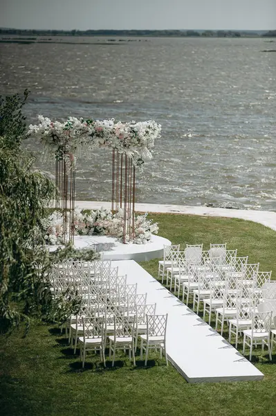 Göl kenarında beyazlarla süslenmiş lüks bir açık hava düğün töreni. Zarif bir çiçek kemeri, şeffaf sandalyeler, yeşil bir çimen ve fevkalade bir arka plan.