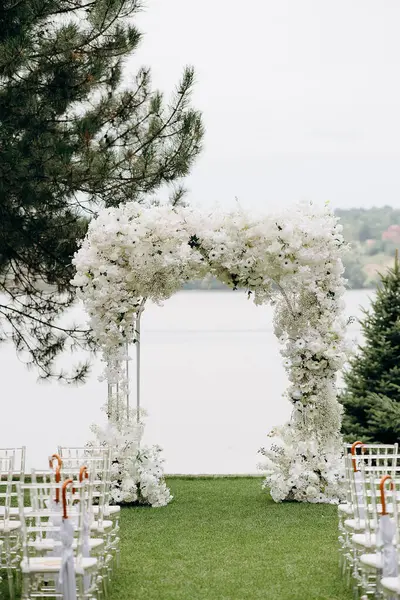 Göl kenarında beyazlarla süslenmiş lüks bir açık hava düğün töreni. Zarif bir çiçek kemeri, şeffaf sandalyeler, yeşil bir çimen, fevkalade doğal bir arka plan.