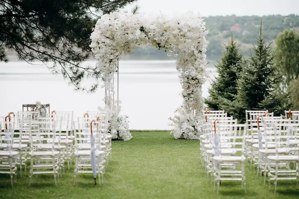 Göl kenarında beyazlarla süslenmiş lüks bir açık hava düğün töreni. Zarif bir çiçek kemeri, şeffaf sandalyeler, yeşil bir çimen, fevkalade doğal bir arka plan.
