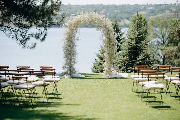 Göl kenarında beyazlarla süslenmiş lüks bir açık hava düğün töreni. Zarif bir çiçek kemeri, yastıklı ahşap sandalyeler, yeşil bir çimen, pitoresk bir doğal.