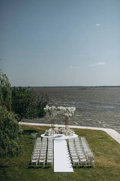 Göl kenarında beyazlarla süslenmiş lüks bir açık hava düğün töreni. Zarif bir çiçek kemeri, şeffaf sandalyeler, yeşil bir çimen ve doğal bir resim.