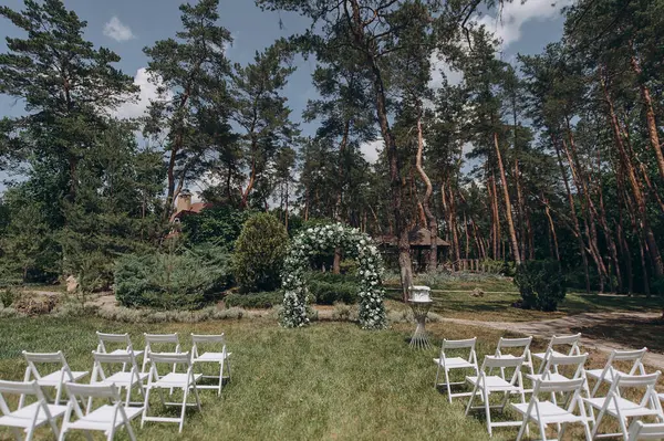 Çam ormanının önündeki bir çayırda düzenlenen düğün töreninde beyaz bir çiçek, yeşil kemer ve zarif beyaz katlanır sandalyeler yer alıyor.