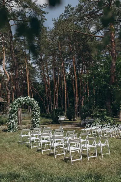 Çam ormanının önündeki bir çayırda düzenlenen düğün töreninde beyaz bir çiçek, yeşil kemer ve zarif beyaz katlanır sandalyeler yer alıyor.