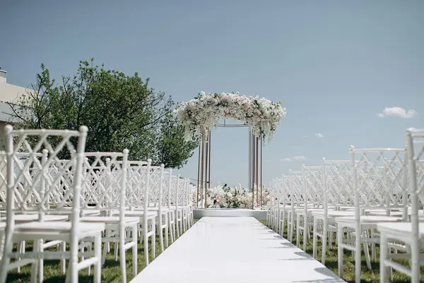 Lüks bir düğün sahnesi, beyaz bir patika boyunca dizilmiş beyaz dantel sandalyeli yüksek çiçekli bir kemer.