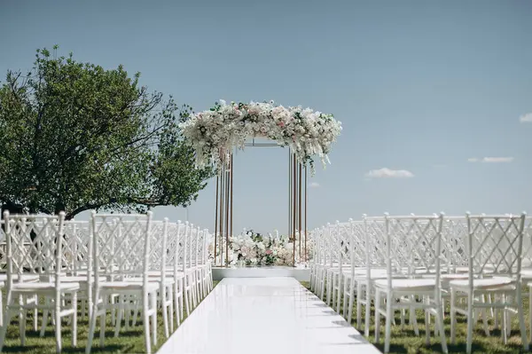 Lüks bir düğün sahnesi, beyaz bir patika boyunca dizilmiş beyaz dantel sandalyeli yüksek çiçekli bir kemer.