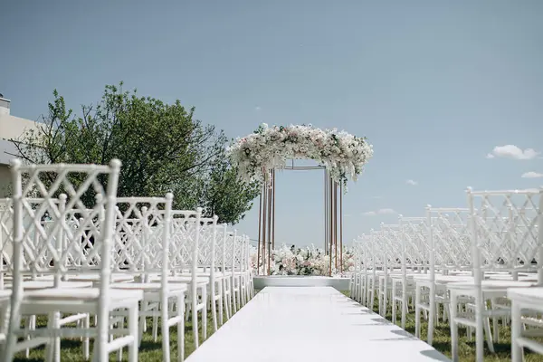 Lüks bir düğün sahnesi, beyaz bir patika boyunca dizilmiş beyaz dantel sandalyeli yüksek çiçekli bir kemer.