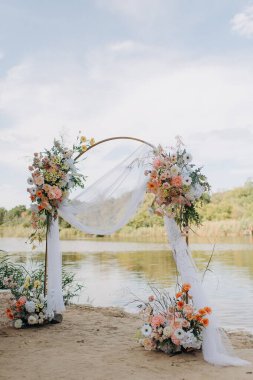 Çiçekli, beyaz kumaşlı bir düğün kemeri sakin bir gölün kumlu kıyısında duruyor.