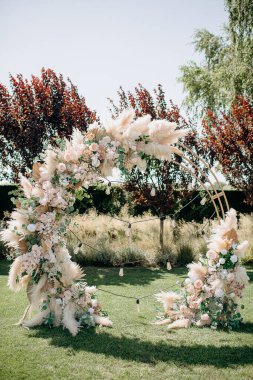 Çiçek aranjmanları ve otlarla süslenmiş dairesel bir düğün kemeri ağaçların ve sicim ışıklarının arasında yeşil bir çimenlikte duruyor ve bohem bir tören yaratıyor.