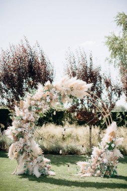 Çiçek aranjmanları ve otlarla süslenmiş dairesel bir düğün kemeri ağaçların ve sicim ışıklarının arasında yeşil bir çimenlikte duruyor ve bohem bir tören yaratıyor.