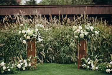 Beyaz çiçek aranjmanlı iki ahşap kaide açık havada uzun otların arasında duruyor.