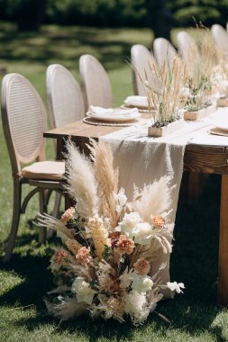 Örülmüş sırtı olan ahşap sandalyeler açık havada yeşil bir çimenlikte otlarla süslenmiş kırsal bir masa hizasında.