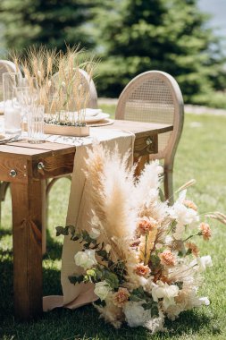 Örülmüş sırtı olan ahşap sandalyeler açık havada yeşil bir çimenlikte otlarla süslenmiş kırsal bir masa hizasında.