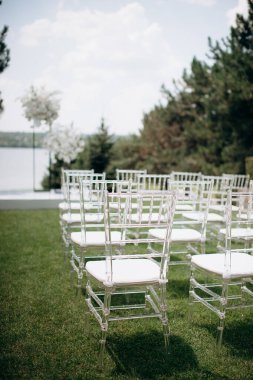 Beyaz yastıklı sıra sıra sandalyeler açık havada bir düğün ortamında göle bakan yeşil bir çimenlikte duruyor.