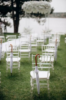 Beyaz yastıklı ve beyaz şemsiyeli sıra sıra berrak sandalyeler açık hava etkinliğinde yeşil bir çimenlikte duruyor.