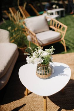 Çiçek aranjmanlı beyaz bir masa bahçedeki yeşil bir çimenlikteki mobilyaların arasında duruyor.