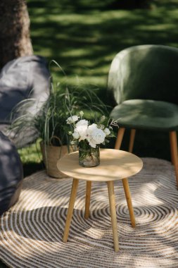 İki yeşil kadife sandalye ve çiçek aranjmanlı küçük bir masa. Bahçedeki çizgili bir halıda duruyorlar.