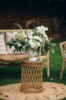 Bir vazonun içindeki beyaz bir çiçek aranjmanı, bahçedeki mobilyaların arasında dokuma bir masanın üzerinde duruyor.