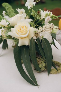 Beyaz ve yeşil çiçekli bir süs, açık hava ortamında beyaz bir masa örtüsünün üzerindeki vazoda limonlar var.