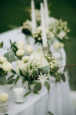 Açık hava etkinliğinde beyaz masa örtüsü üzerinde mumlar olan beyaz bir çiçek süsü.