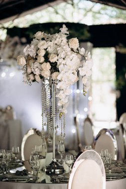 Beyaz gülleri ve orkideleri olan uzun bir çiçek süsü bir etkinliğin ortasında aynalı bir masada duruyor.