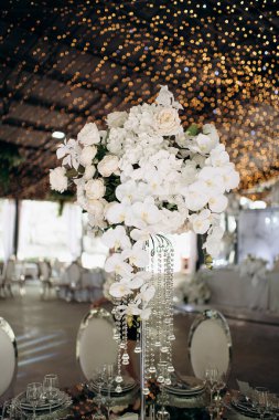 Beyaz gülleri ve orkideleri olan uzun bir çiçek süsü bir etkinliğin ortasında aynalı bir masada duruyor.