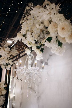 Bir kristal avize, kapalı bir alanda, beyaz bir çiçekliğin altında asılıdır. Koyu tavanı ve parlak ışıkları vardır.