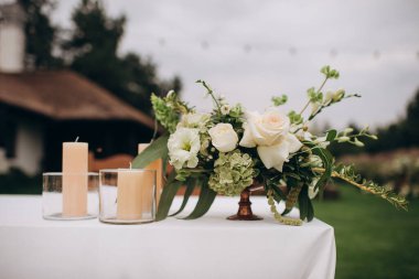 Zarif bir açık hava sofrasında, bahçesinde güller, bahçeler, okaliptüsler ve bronz bir vazoda yeşil aksanlı beyaz bir çiçek süsü bulunur.