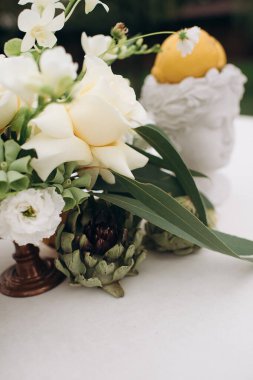 Beyaz bir masa örtüsü üzerinde güller, ranunculus, orkideler ve enginarlar bulunan bir çiçek aranjmanı yakalar. Bir limon kafa şeklindeki bir terracotta tenceresinde oturur ve canlı bir aksan ekler. Okaliptüs yaprakları doğal zarafeti vurgular