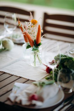 Açık bir masadaki beyaz vazoda havuç ve sebzelerle çiçek aranjmanı.