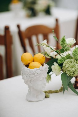 Beyaz güller ve beyaz bir masa örtüsünün üstündeki vazoda limonlar olan zarif bir masa.