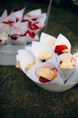 Kırmızı ve sarı gül yapraklarıyla dolu beyaz kağıt koniler çimlerin üzerinde beyaz bir kasede