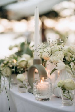 Açık hava ziyafeti sırasında gün batımı, beyaz lavanta gölgeli çiçeklerin ve cam vazolarda çeşitli mumların bulunduğu bir kompozisyonun yakın çekimi, romantik ve zarif bir atmosfer yaratıyor.