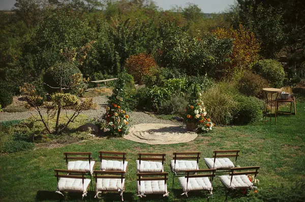Avluda bir çayır, düşük çiçekli bir kemer ve büyük ahşap sandalyelerle bir düğün töreni düzenlenir, kırsal ve samimi bir açık hava ortamı yaratır.