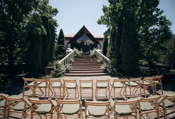 Taştan bir terasta sıralanmış ahşap sandalyeler yemyeşil yaprakların arasında bir bahçeye çıkıyor.