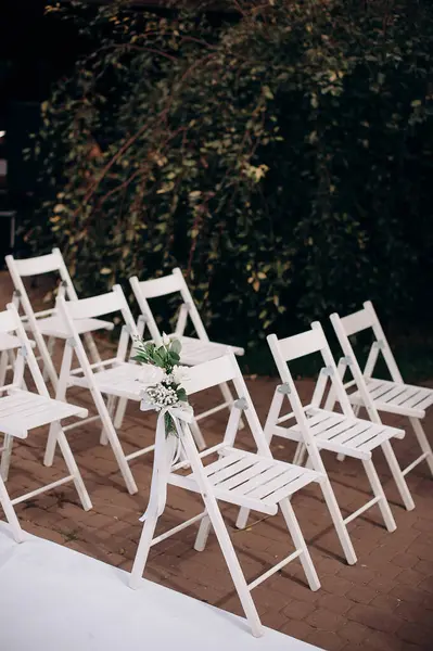 Çiçek buketleriyle süslenmiş beyaz ahşap sandalyeler açık havada bir taş terasta dizilmiş.