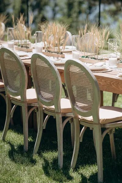 Örülmüş sırtı olan ahşap sandalyeler açık havada yeşil bir çimenlikte otlarla süslenmiş kırsal bir masa hizasında.