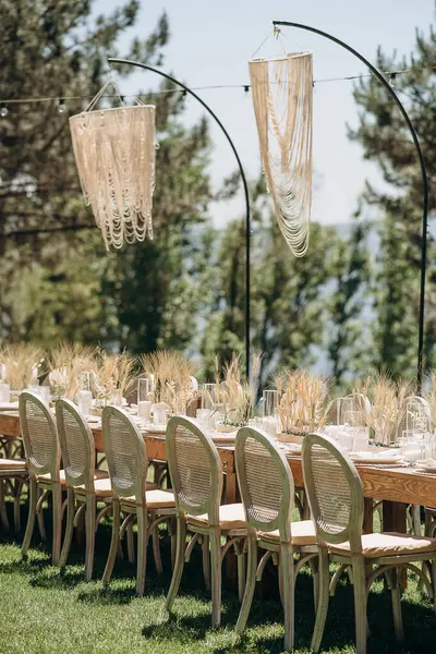 Örülmüş sırtı olan ahşap sandalyeler açık havada yeşil bir çimenlikte otlarla süslenmiş kırsal bir masa hizasında.