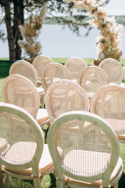 Yuvarlak dokuma sandalyeler, arka planda bir göl bulunan yeşil çimenlikteki çiçek kemerine bakıyor.