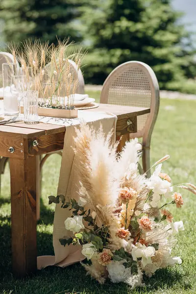 Örülmüş sırtı olan ahşap sandalyeler açık havada yeşil bir çimenlikte otlarla süslenmiş kırsal bir masa hizasında.